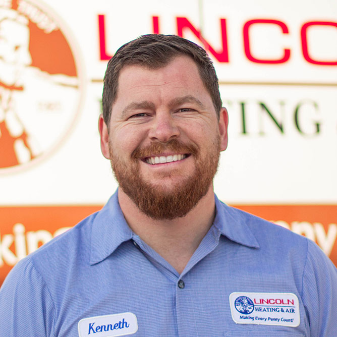Kenneth Q., General Manager at Lincoln Heating & Air, smiling in a blue work shirt with a name tag, in front of a company banner emphasizing family-oriented heating and air conditioning services.