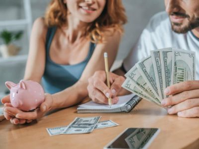 Couple discussing finances with a piggy bank, cash, and a notebook on a wooden table, illustrating budgeting and cost-saving for heating services.