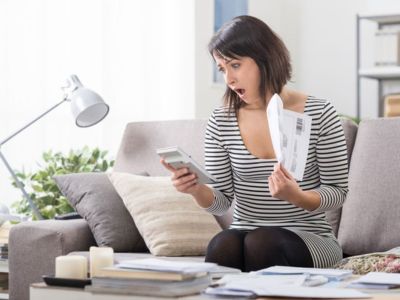 Woman sitting on a couch, shocked while reviewing high energy bills on a calculator and holding a bill in hand, illustrating concerns about heating efficiency and rising utility costs related to HVAC maintenance.