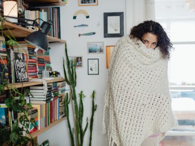 Woman wrapped in a cozy blanket, appearing cold in a home library setting with bookshelves and plants, symbolizing the need for efficient heating during winter months.