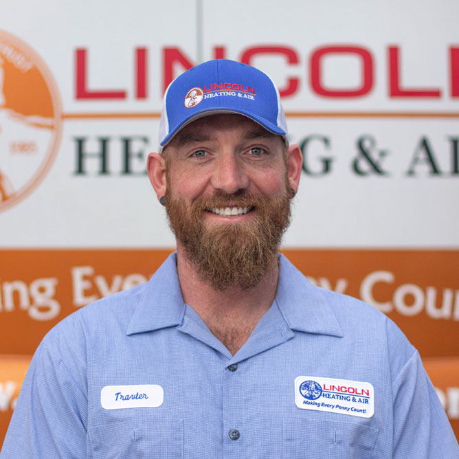 Service technician Travler G. smiling, wearing a blue uniform and cap with Lincoln Heating & Air logo, in front of company branding.